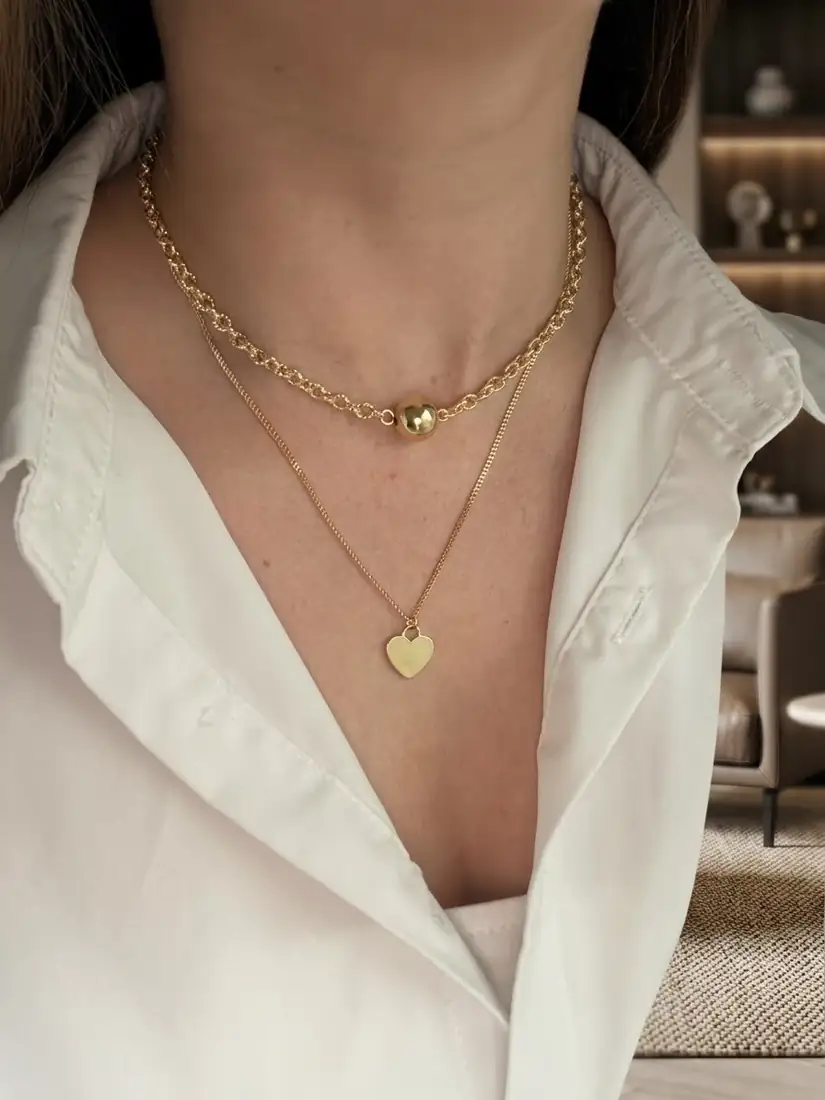 Dos collares en tono dorado sobre una camisa blanca: una cadena con una esfera central y otra más fina con un colgante de corazón.
